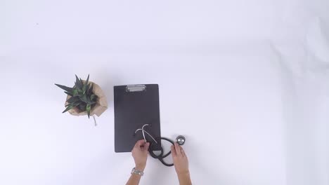 top-down view of medical items placed on white background with a stethoscope, syringe, black dossier folder, white glasses, and small plants