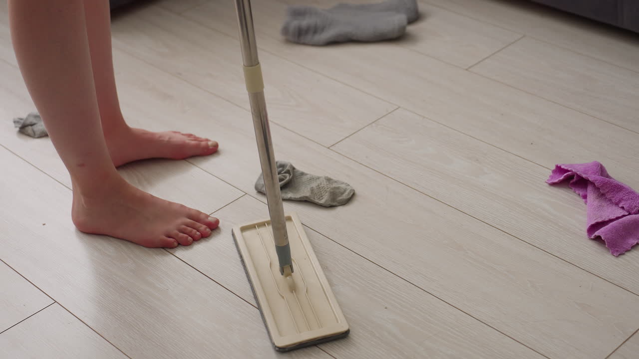 Close up of person legs walking barefoot on tiled floor holding mop, scattered socks and clothes lying around near sofa in messy living room illuminated by sunlight through curtains