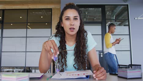 mujer de negocios creativa biracial en el escritorio, leyendo, tomando notas y hablando durante una videollamada