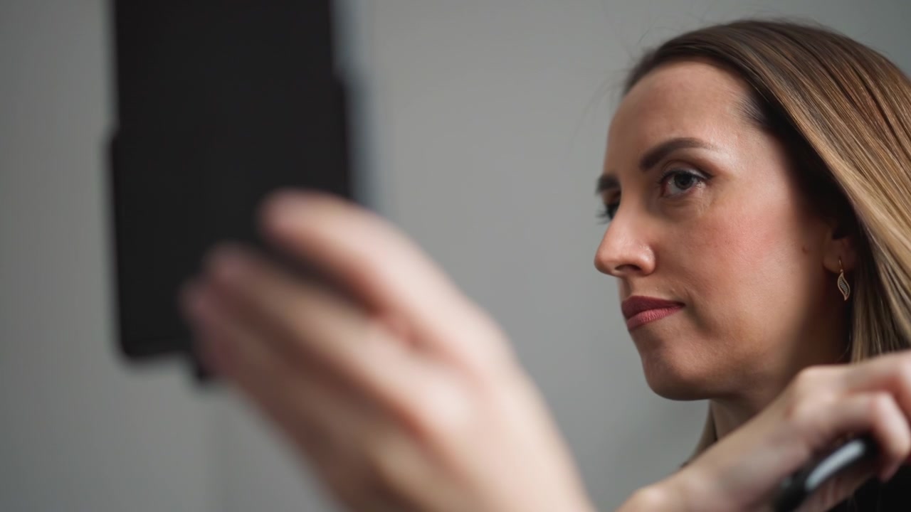 Primo piano di una donna che si spazzola i capelli davanti allo specchio, con attenzione all'azione e ai dettagli. Il soggetto tiene un oggetto vicino al viso, con lunghi capelli castani lisci e indossa abiti scuri