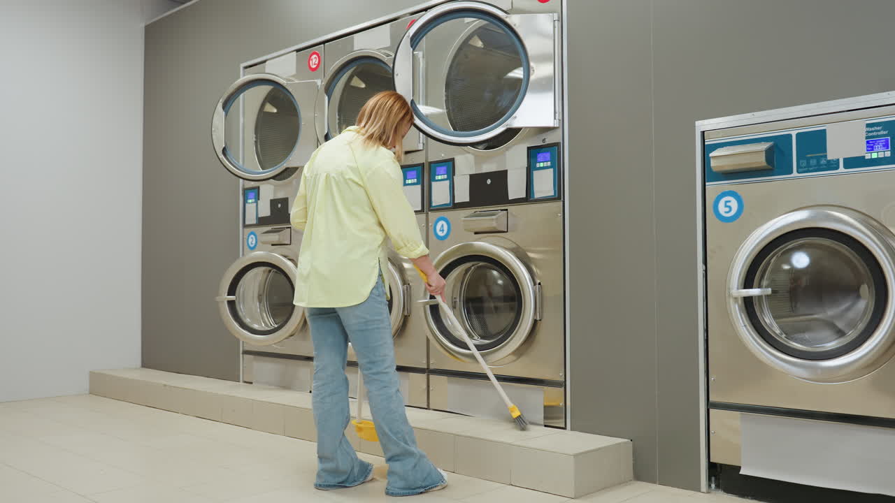 Back view casual woman with broom and dustpan moves toward industrial washing machines in laundromat, sweeping floor near stainless doors, showing routine cleaning and quiet service environment