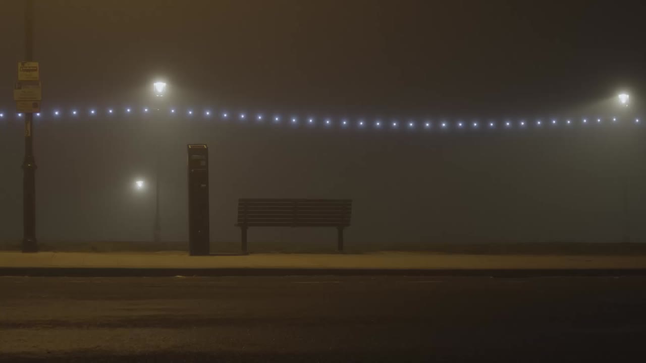Dead of night, quiet road, illuminated bench and a parking meter under a string of diffused street lights in the fog. North east England. Cinematic mysterious and eerie, shot in slomo.