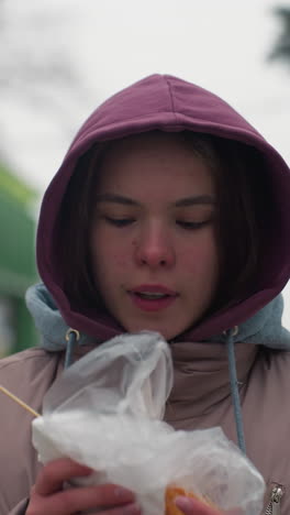 mujer joven con chaqueta de invierno comiendo pasteles al aire libre en un parque urbano, sosteniendo comida envuelta, clima frío, estilo de vida casual, capucha, disfrutando de la comida callejera en la temporada de invierno