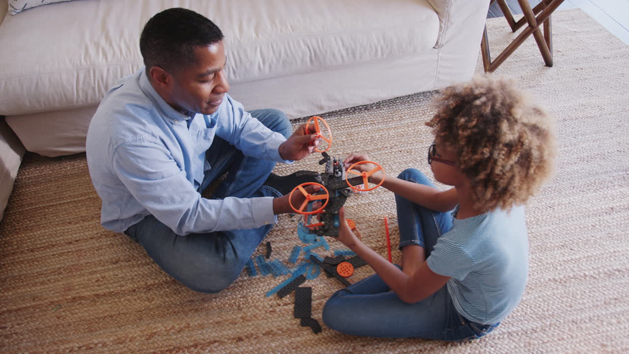 Pre-teen African American girl and grandad sitting cross legged on the floor constructing a toy, elevated view