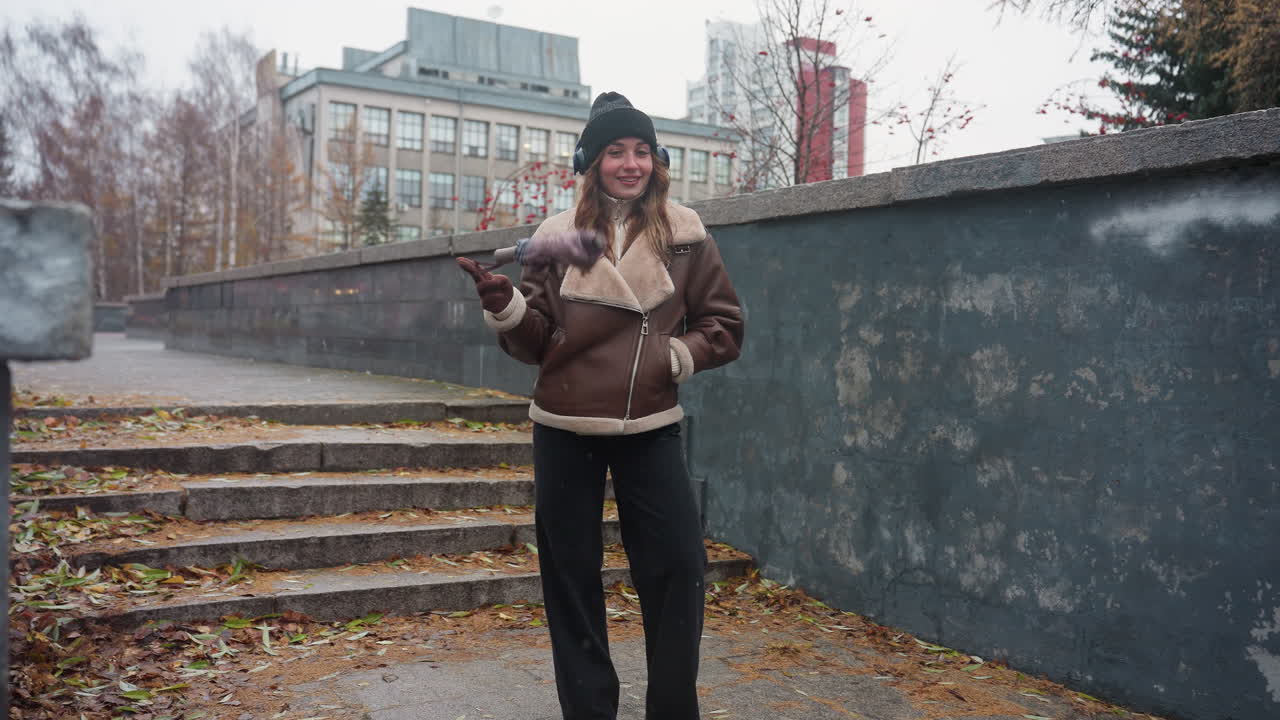 Young girl in black knit cap, brown shearling jacket, black trousers stands on outdoor concrete path with autumn leaves, swinging umbrella playfully with one hand in pocket under light snowfall
