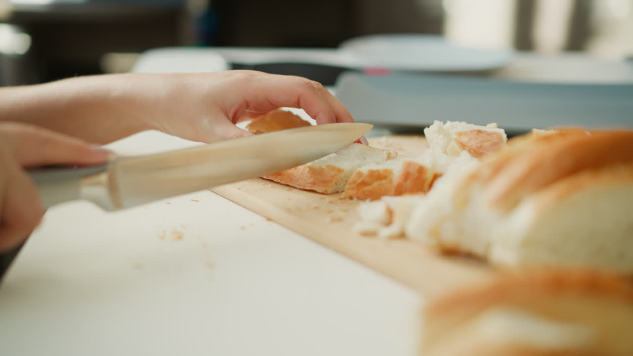 Soft window light highlights side view of griller hands slicing loaf into small bite sized pieces on white table with scattered crumbs, capturing focused calm culinary preparation process