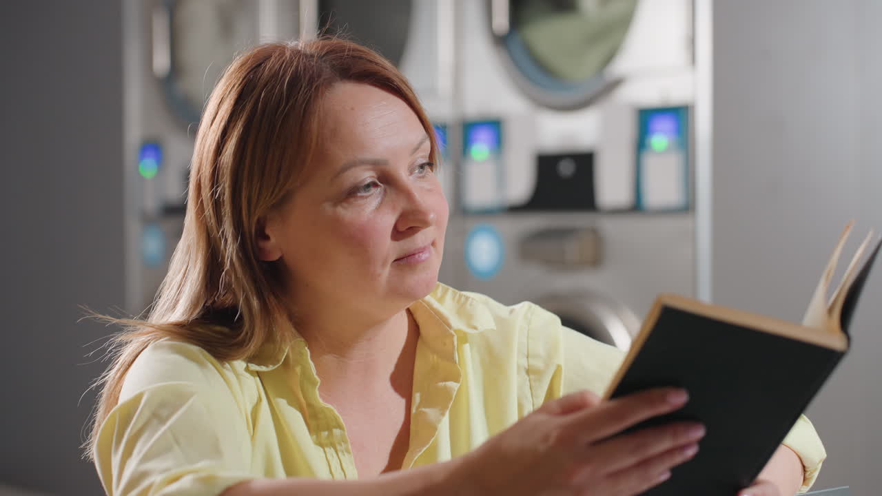 Laundry manager seated inside laundromat opens record book, checking notes while holding coffee cup on shiny table, industrial washers with digital panels glow in background