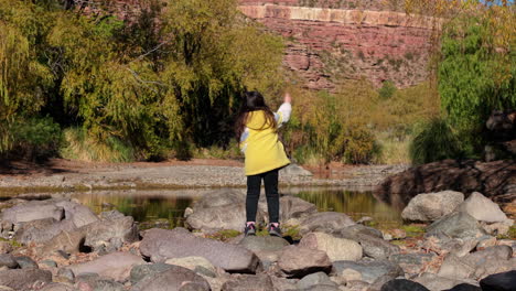a young child in yellow jacket throwing a stone into the calm lake in a rocky nature scene.