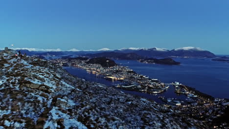 aerial view over a snowy mountain, revealing the alesund city, blue hour in norway