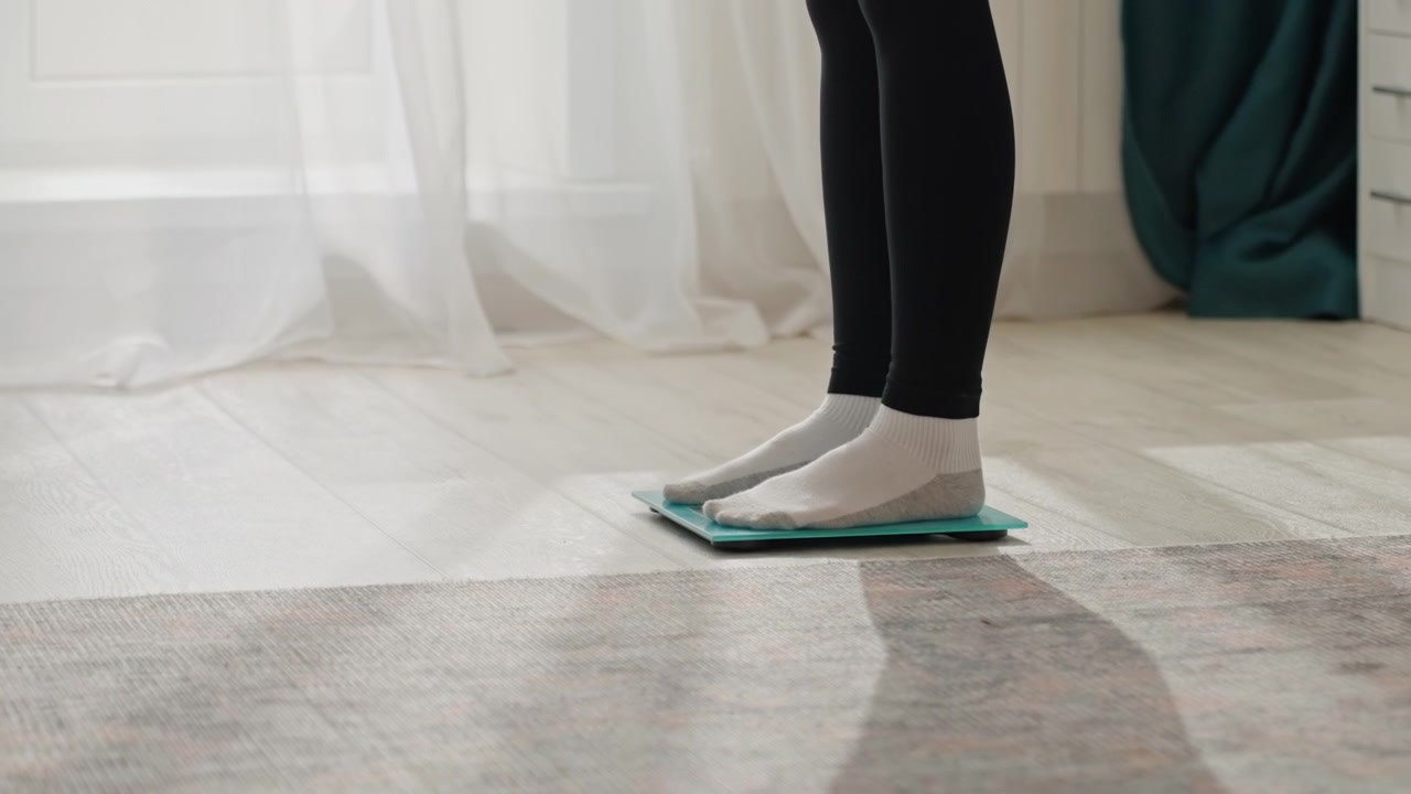 Home bedroom sockson feet testing scale. Soft morning light through sheer curtains, teal glass scale on rug, socksclad feet step tentatively onto digital display minimal interior, calm mood, focus
