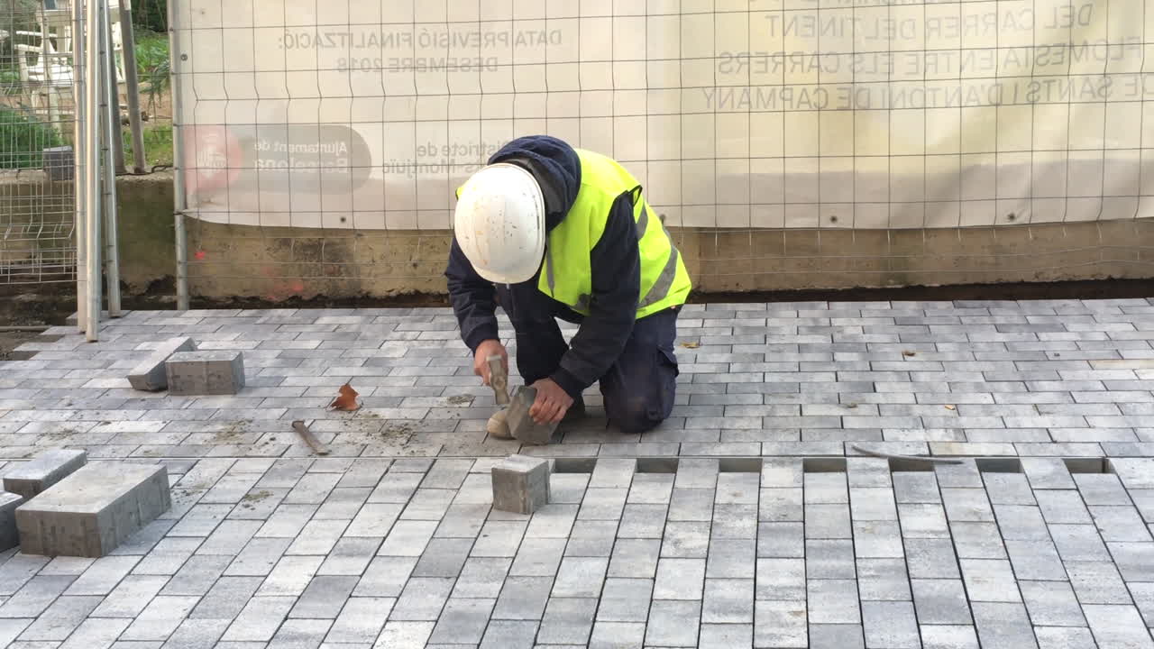 Public Ajuntament de Barcelona worker with hammer, helmet and yellow jacket hammering a square block of concrete on under construction pedestrian street or sidewalk