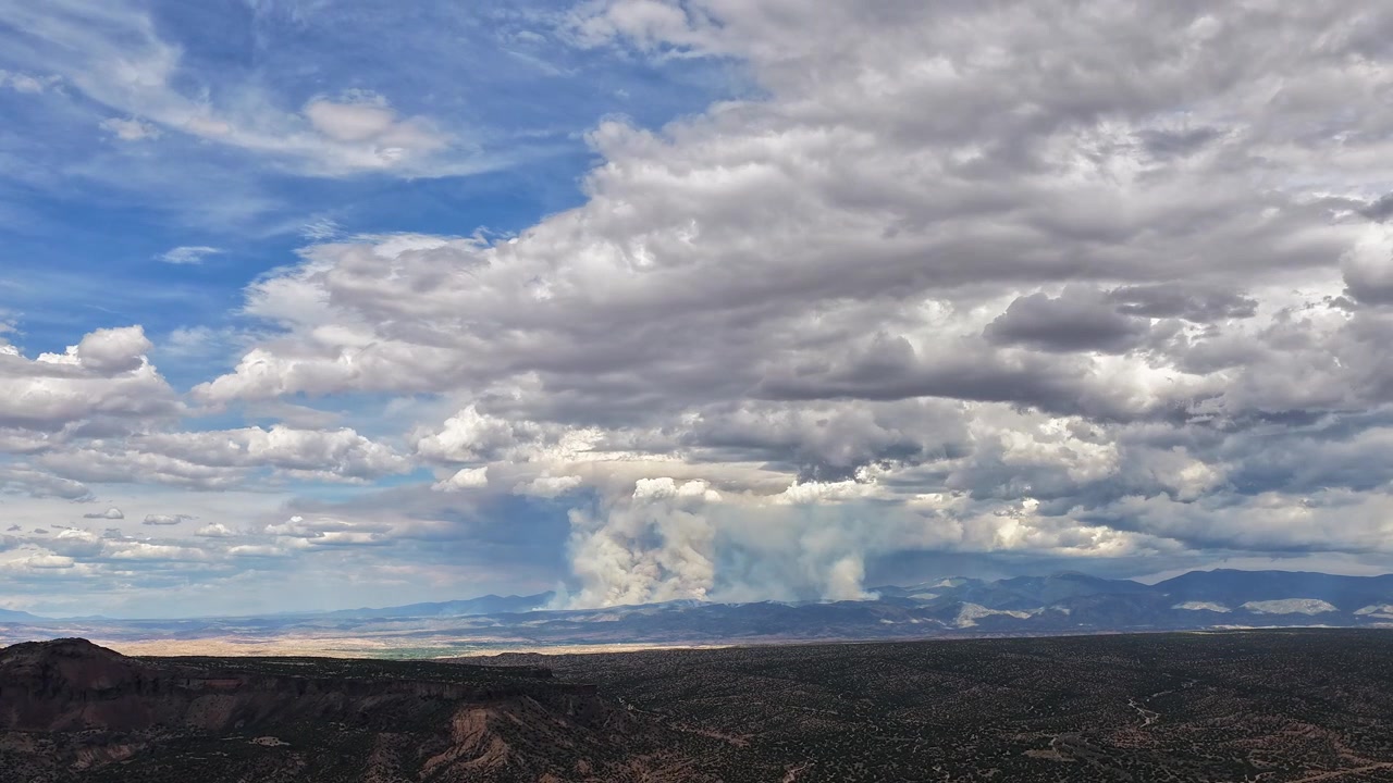 Timelapse 4K con drone del massiccio incendio boschivo di Frijoles che brucia nella contea di Santa Fe, NM. Un'imponente colonna di fumo si innalza dal fianco della montagna distante sotto un cielo drammatico e mutevole di nuvole pesanti.