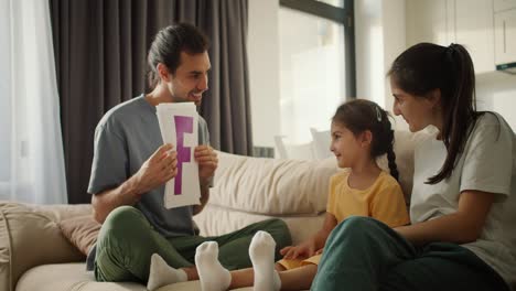 A-brunette-man-in-a-gray-T-shirt-teaches-his-little-daughter-in-a-yellow-dress-various-letters,-showing-them-on-pieces-of-paper.-Little-girl's-parents-help-her-learn-different-letters-by-showing-colorful-letters-on-a-sheet-of-paper