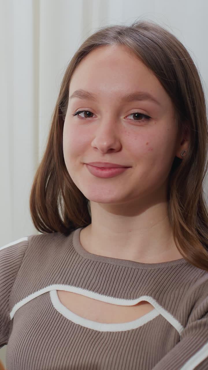 joven leyendo un libro de portada marrón en un entorno pacífico junto a una ventana con cortina transparente, sonríe cálidamente mientras hojea las páginas, disfrutando de un momento tranquilo y relajante
