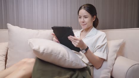 beautiful asian woman comfortably sit on the couch using smart pen touching tablet searching for online store shop, online customer shopping, home convenience, trading e-commerce online purchasing