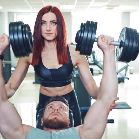 beautiful trainer woman helps a young man lifting heavy dumbbells. sporty man lifts dumbbells while lying on special desk in the gym. personal trainer and a bodybuilder.