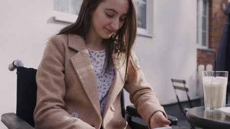 feliz niña discapacitada en silla de ruedas usando teléfono móvil en una terraza de bar