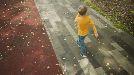 aerial rear view of kid in yellow top walking along patterned tiled path covered with fallen autumn leaves, arms raised playfully while exploring outdoor park area surrounded by natural foliage