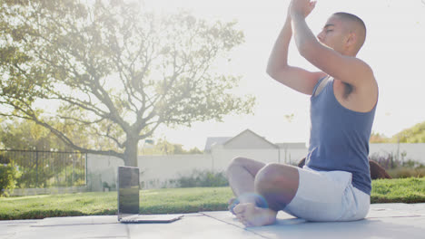 Fokussierter-Biracial-Mann,-Der-Yoga-Mit-Laptop-Im-Sonnigen-Garten-Praktiziert,-Zeitlupe