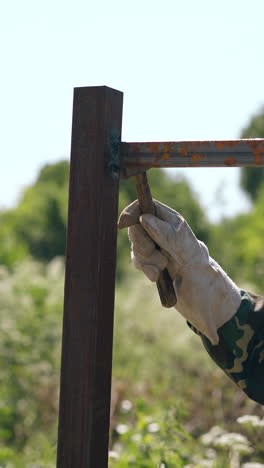 mature handyman in work gloves taps with hummer on fixed fence carcass details against weeds