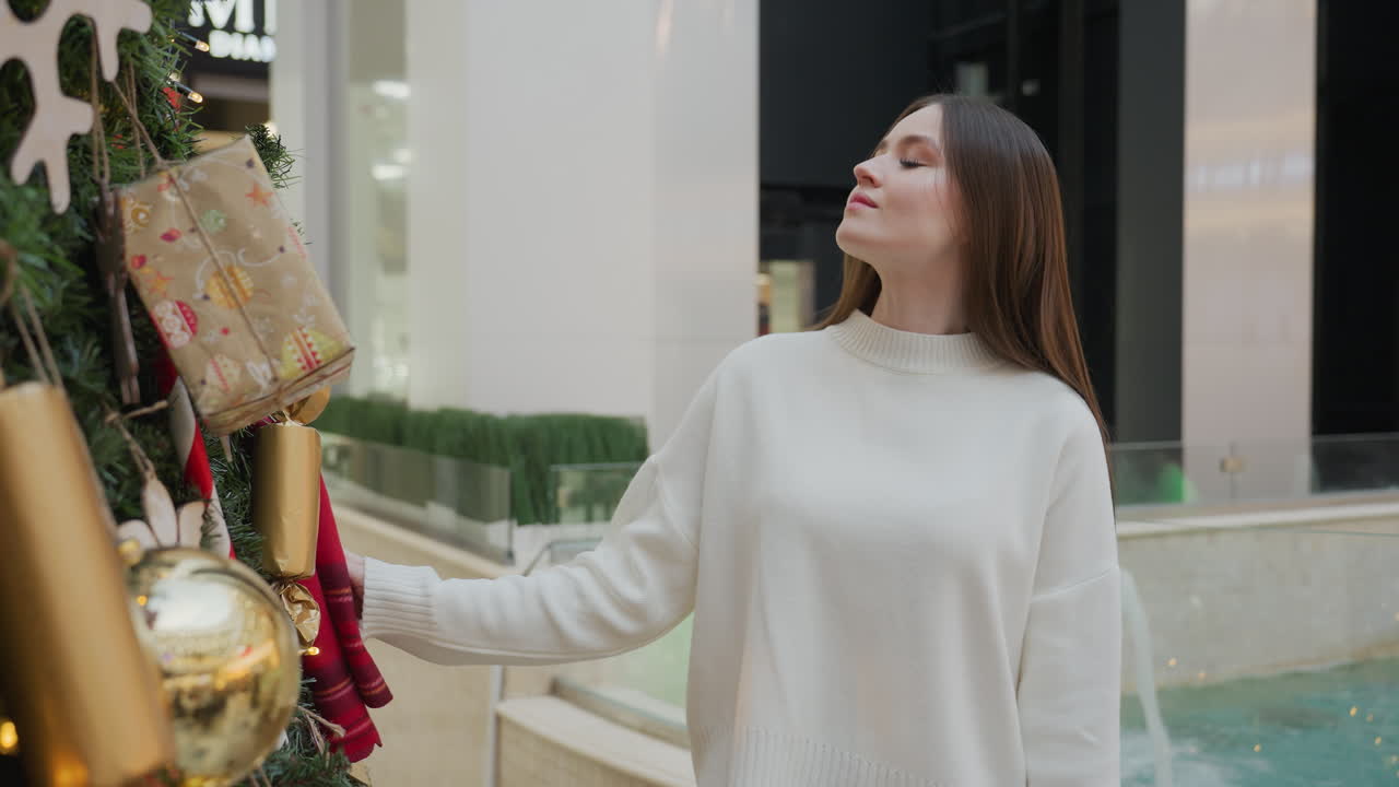 Woman in white sweater admires Christmas tree with glass-bordered water spring and decorative plant, urban structures in background, showcasing holiday ambiance in shopping mall