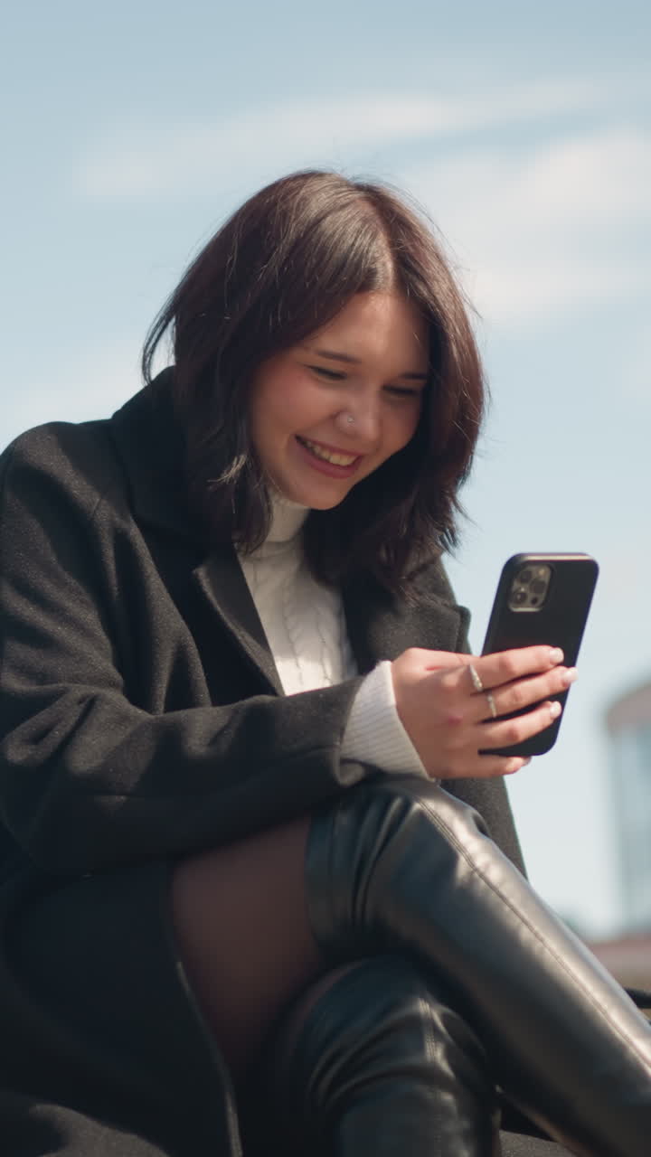 una joven blanca alegre sonriendo mientras se centra en su teléfono inteligente en un parque con las piernas cruzadas, un fondo borroso con un edificio y hierba verde, disfrutando de su momento al aire libre