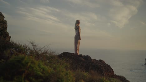 charming blond woman standing on cliff edge spinning head swaying hair, morning sunlight