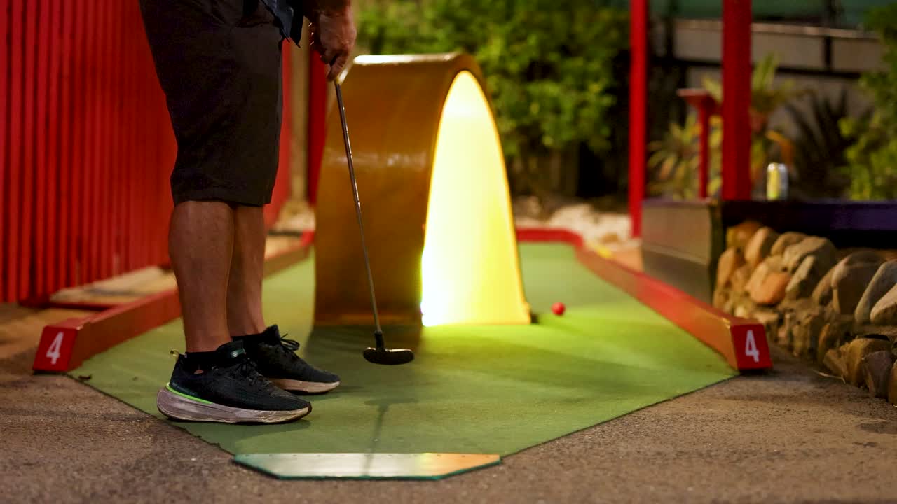 A person attempts a putt on a mini golf course at night, illuminated by warm lighting, creating a playful atmosphere