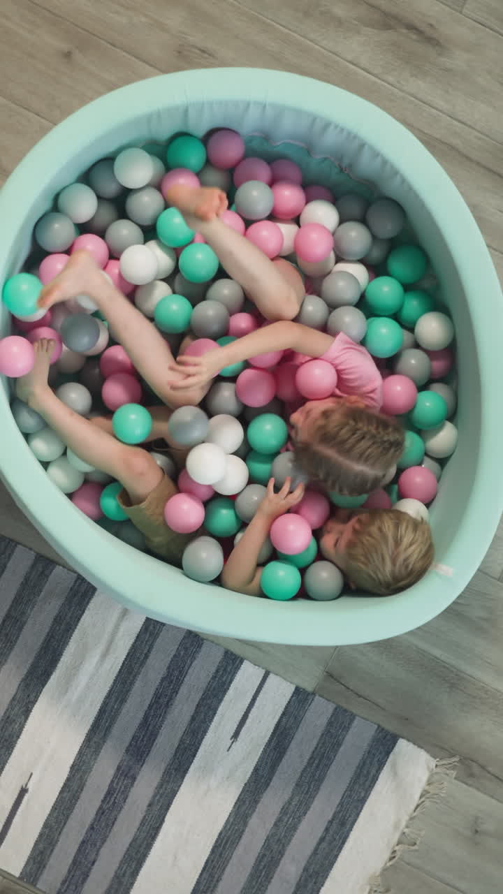 Amused little girl with younger brother lie in dry pool playing with white, pink, gray and light blue plastic balls. Cheerful kids siblings rest at home