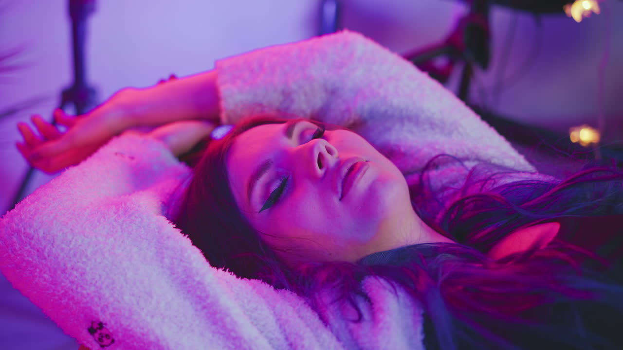 Woman peacefully sleeping with hands stretched above head while lying under vivid pink and purple lights, wearing fluffy sweater, surrounded by blurred studio background with soft dreamy atmosphere