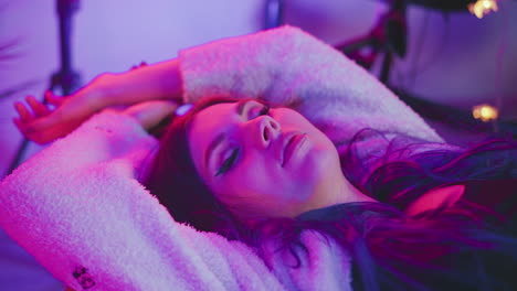 woman peacefully sleeping with hands stretched above head while lying under vivid pink and purple lights, wearing fluffy sweater, surrounded by blurred studio background with soft dreamy atmosphere
