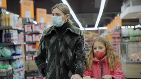 mom and daughter roll a shopping cart in front of them. they walk between the rows of goods in the supermarket and choose what to buy.
