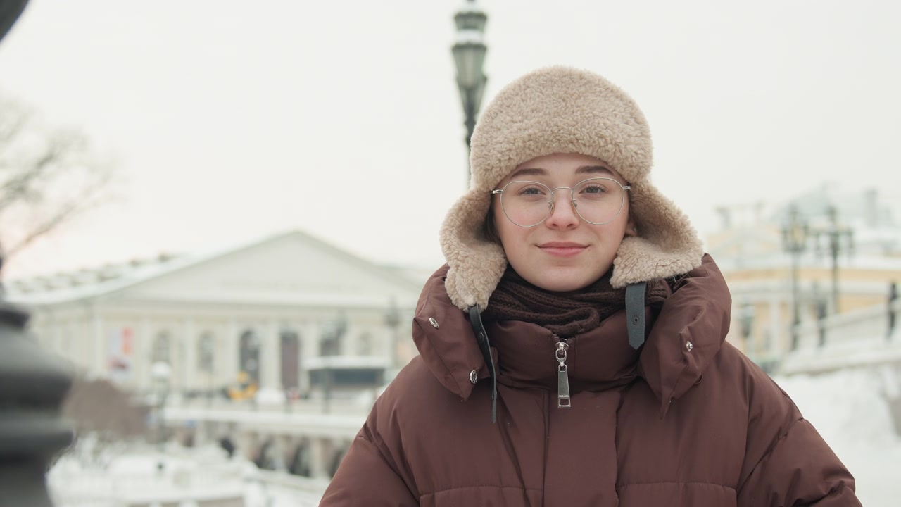 Una giovane donna in una piazza urbana innevata allaccia un cappotto imbottito marrone e sistema un cappello di maglia beige con paraorecchie, in piedi con calma con occhiali trasparenti in un ambiente stradale invernale e una luce soffusa e nuvolosa.