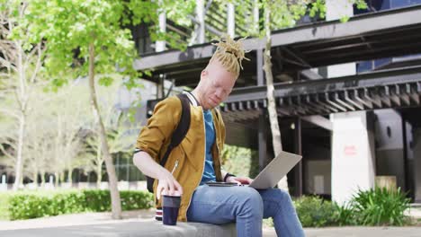 thoughtful albino african american man with dreadlocks sitting in park drinking coffee, using laptop