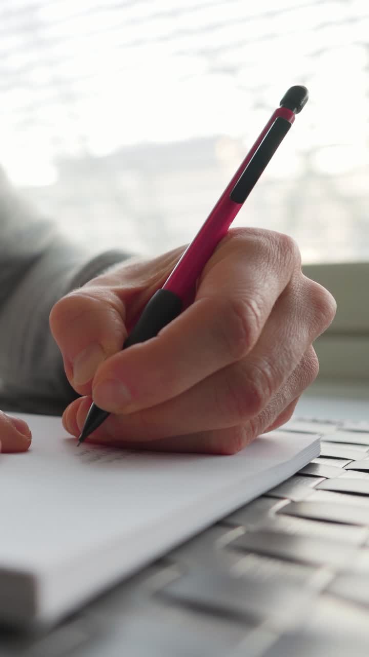 Left-handed man writing on paper next to a window