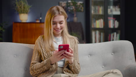 Mujer-Joven-Feliz-Charlando-En-El-Teléfono-Inteligente-Y-Riendo-Mientras-Se-Sienta-En-Un-Sofá-En-Casa