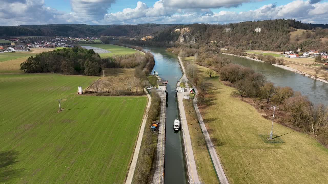 This footage shows a boat on a German canal, situated between lush fields and a dense forest, with a lock system visible in the distance