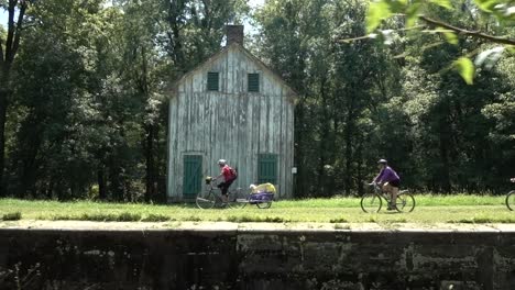 mature woman pulling a trailer with a dog in it cycles past an old white wooden building on the c-o canal national historic park near harpers ferry, west virginia