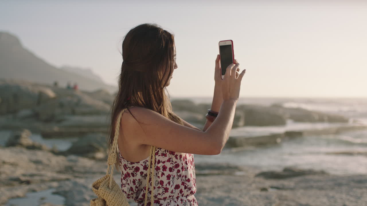 mujer joven tomando una foto usando el teléfono en la playa fotografiando el atardecer