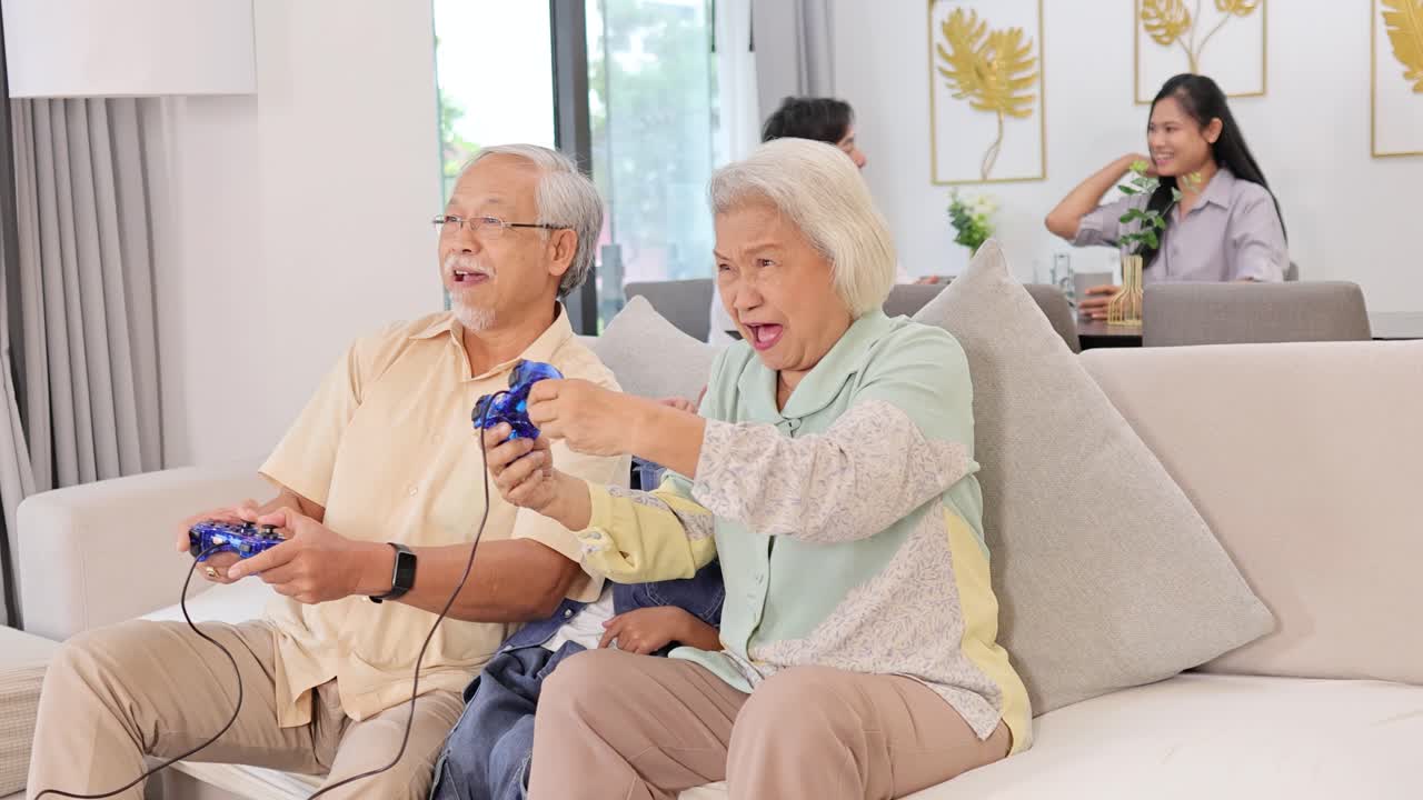 An elderly couple enthusiastically plays a video game on a couch, with family in the background, in a bright living room