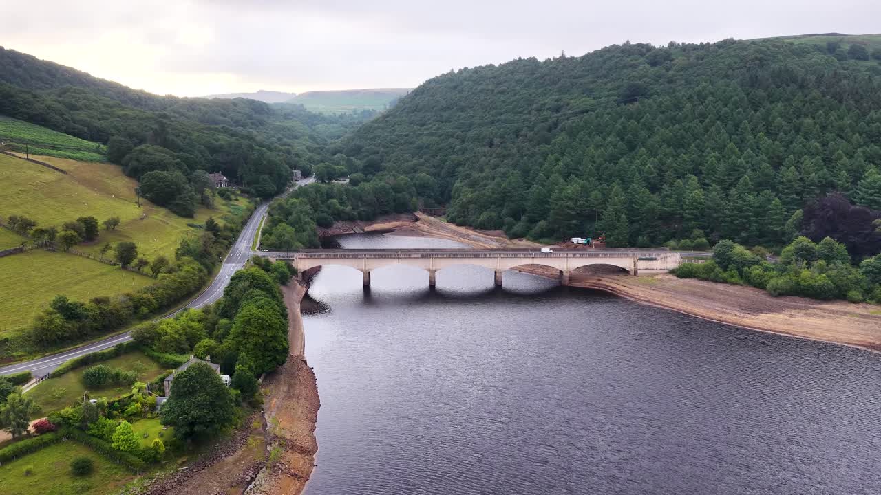 Drone footage glides above an arched stone bridge spanning Ladybower Reservoir, surrounded by lush green hills and forest under soft daylight