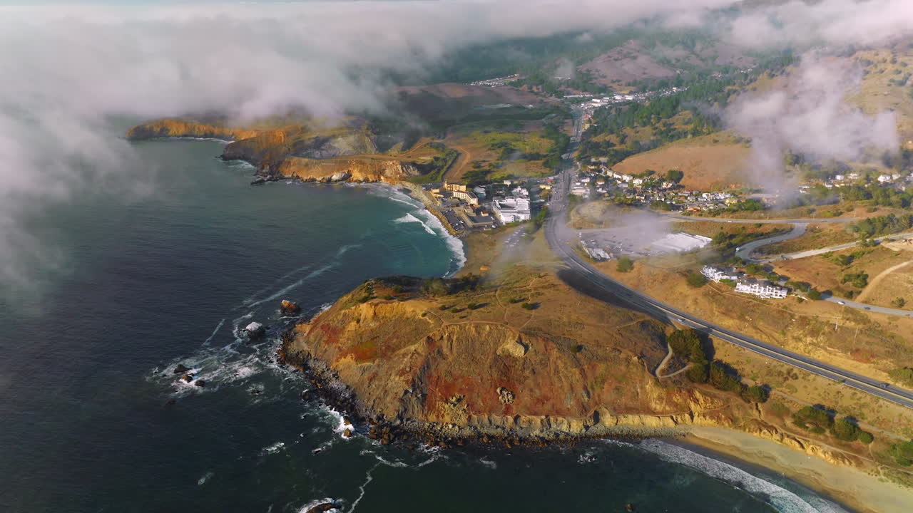 Sunny rocky coastline with houses and highway on top. Thick fog crawling to the city. Aerial perspective.
