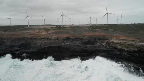 Toma-Panorámica-Aérea-De-Un-Parque-Eólico-Al-Fondo-Con-Agua-Del-Océano-Golpeando-La-Costa-De-Cape-Bridgewater,-Australia