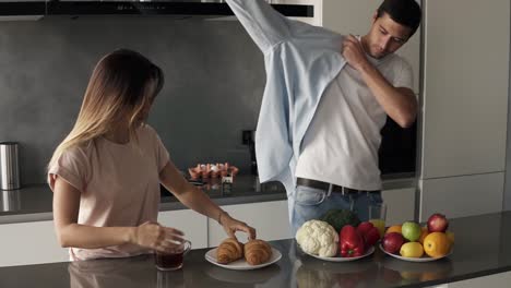 morning at the kitchen counter, fast breakfast before work - croissants and coffee for breakfast. handsome husband came to the kitchen and putting on the shirt, in a hurry, wife eating croissants