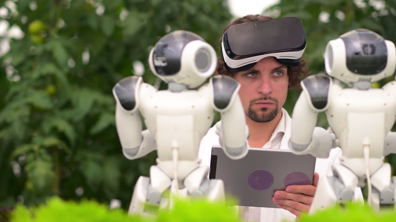 Laboratory technician in a white coat with virtual reality headset holding a tablet while analysing two humanoid robots in a greenhouse farm