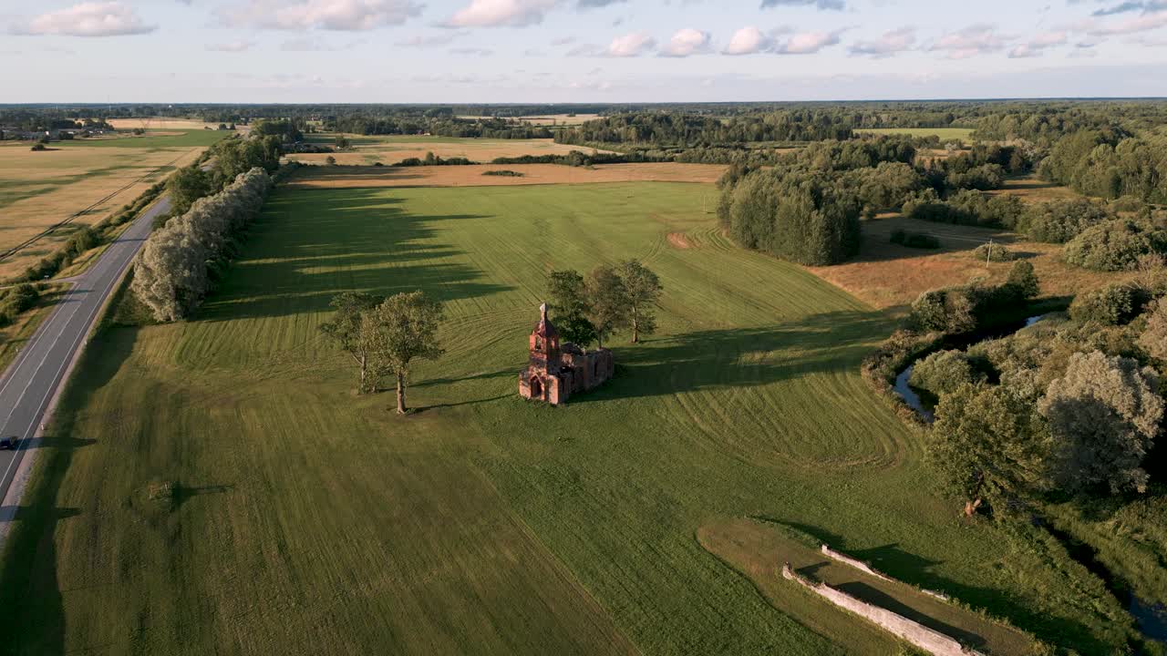 Aerial drone flyover baltic country side of Estonia, Europe with a orthodox or catholic lost place church ruin without roof on a green meadow next to country road with cars on a hot summer day 2022