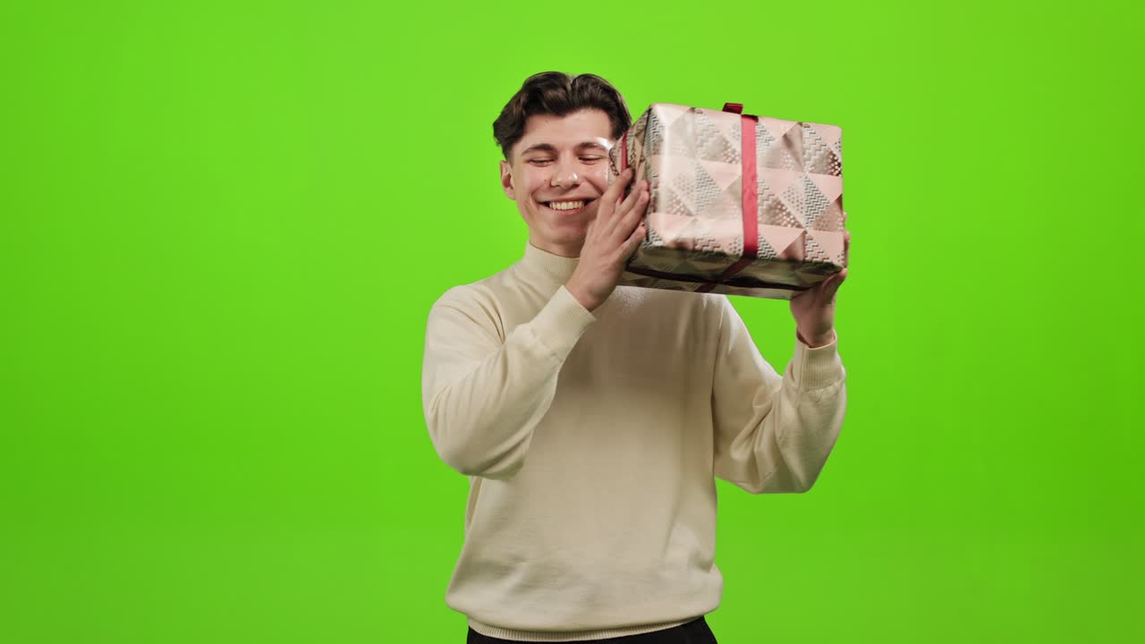 그 남자는 귀 근처에 있는 선물 상자를 흔들고, 그 안에 무엇이 있는지 알고 싶어하고, 미소 짓고, 초록색 바탕에 서 있다.