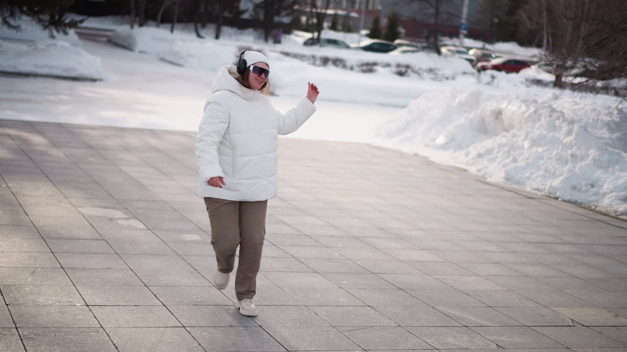 Rear view of woman wearing fluffy white jacket, beanie and headphones dancing energetically on snow dusted urban plaza, playful winter movement captured against backdrop of piled snow and parked cars