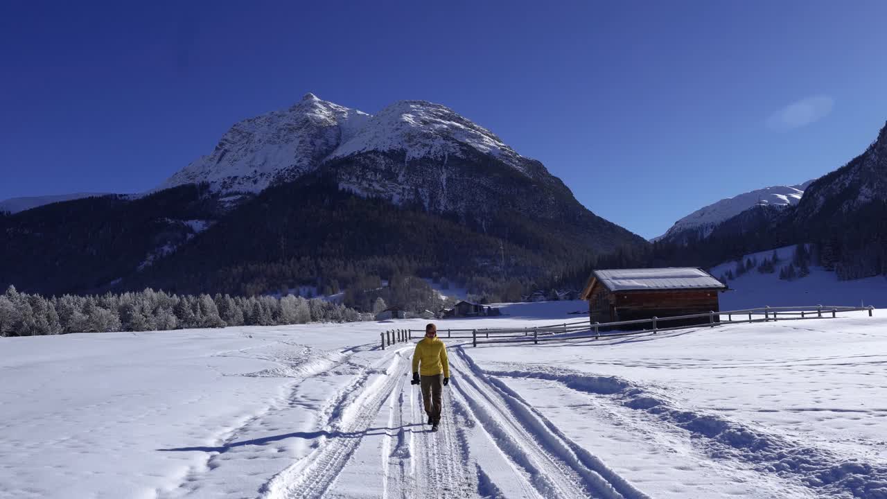 Giovane uomo caucasico con una giacca gialla e un berretto di lana che cammina attraverso un paesaggio invernale innevato con una baita in legno.