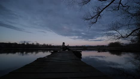 silhouette of musician in black playing guitar sitting on pier embankment on sunset